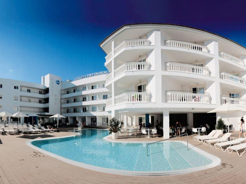 Hotel bianco moderno con grande piscina e lettini sotto un cielo sereno.