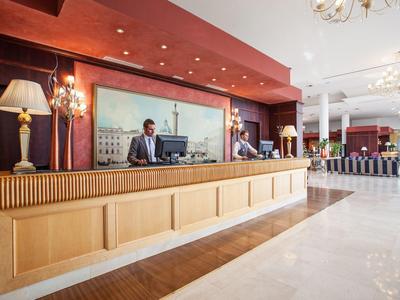 Große Hotelrezeption mit hellem Holz, roter Wand und zwei Mitarbeitern am Tresen.