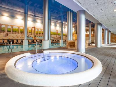 Moderner Whirlpool mit blauer Beleuchtung auf Holzterrasse neben Glasfront und Säulen.