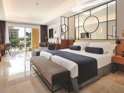 Modern hotel room with large bed, bench, and balcony view of garden.