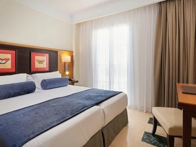 Hell eingerichtetes Schlafzimmer mit zwei Einzelbetten, blauen Akzenten und großem Fenster mit Vorhängen.