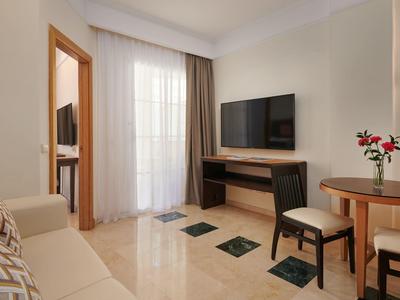 Helles Wohnzimmer mit Sofa, großem Fernseher, Rundertisch, zwei Stühlen und Blumen in Vase.