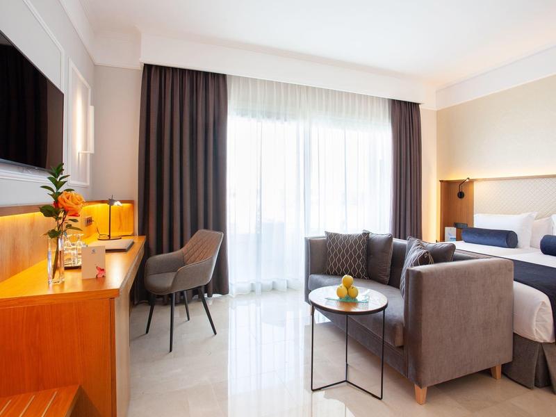 Moderne Hotelzimmer mit großem Bett, Sofa, Tisch, Schreibtisch und TV, heller Raum mit Vorhängen.