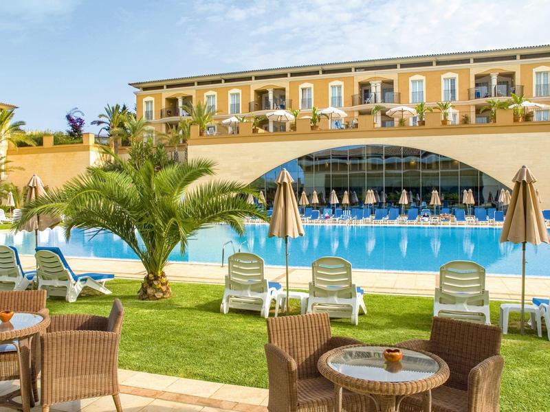 Hotelgarten mit Swimmingpool, Liegestühlen, Sonnenschirmen und mehrstöckigem Gebäude im Hintergrund.