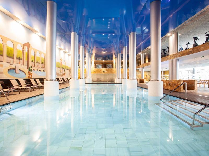 Helles Hallenbad mit Säulen, Liegestühlen und moderner Beleuchtung.