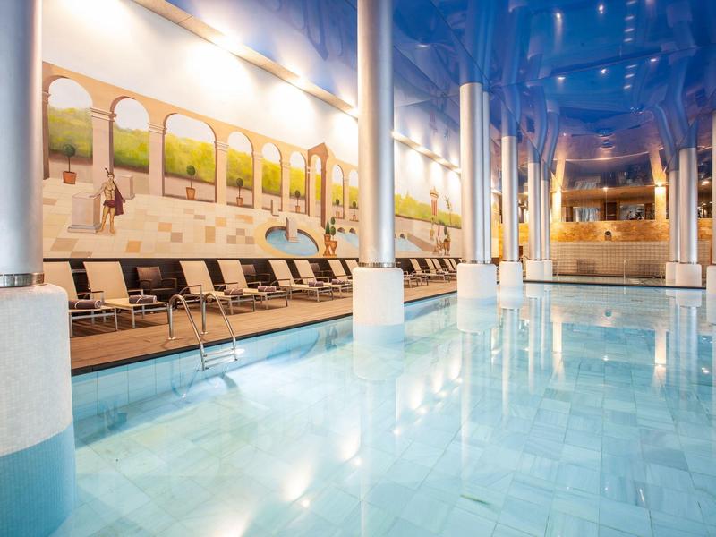 Innenbereich eines hellen Schwimmbads mit Wasser, Säulen, Liegestühlen und Wandbildern.