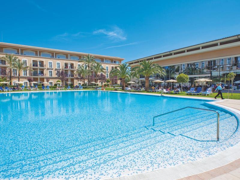 Großer runder Pool vor einem dreistöckigen Hotelgebäude bei klarem blauem Himmel.