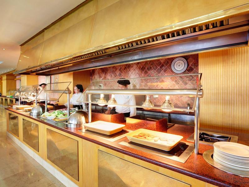 Buffet mit warmen Speisen, Holztheke, Tellerstapel und Küchenpersonal im Hintergrund.