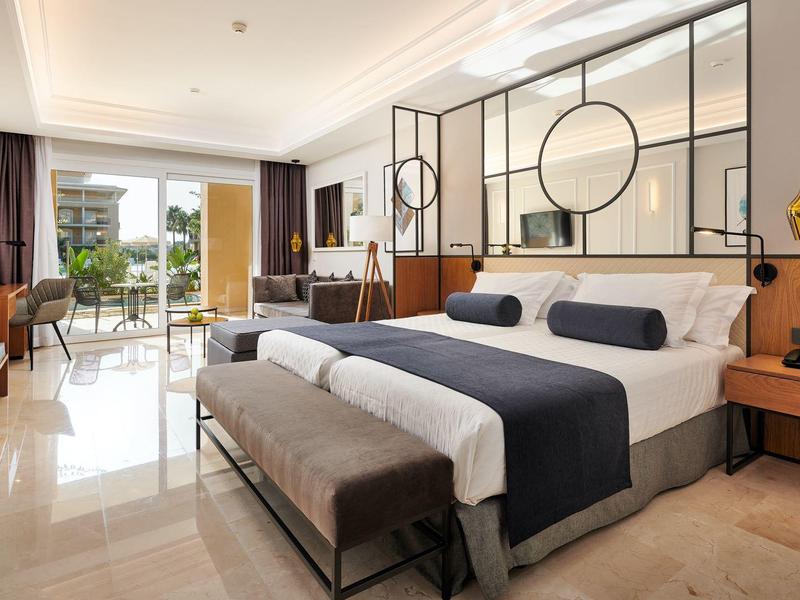 Helles Hotelzimmer mit großem Bett, modernen Möbeln und Blick auf eine Terrasse.