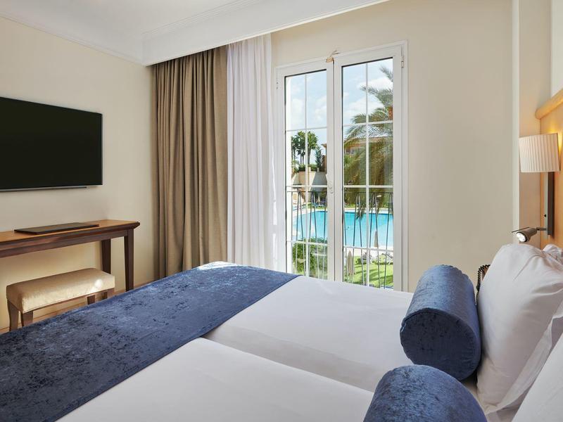 Helles Schlafzimmer mit Doppelbett, blauen Kissen, TV an der Wand und Blick auf Pool durch Glasschiebetür.