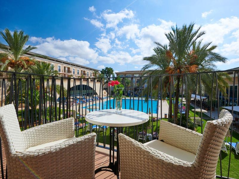 Blick auf einen Pool mit Sonnenschirmen, Palmen und zwei weißen Korbsesseln um einen kleinen runden Tisch.