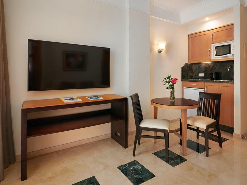 Moderner Wohnraum mit Fernseher, Holztisch, zwei Stühlen und Küchenzeile in neutralen Farben.