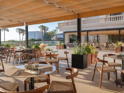 Terraza exterior de hotel con mesas, sillas y plantas bajo un techo sombreado.