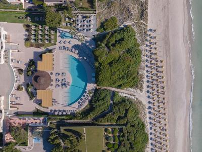 Vista aérea de un hotel frente a la playa con piscina, tumbonas, sombrillas y jardín verde.