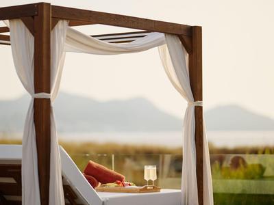 Cama de día acogedora con cortinas y cojines en terraza con vistas a montañas al atardecer.