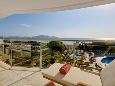 Balcón con tumbonas y barandilla de cristal con vistas a la piscina, jardín y mar bajo cielo despejado.