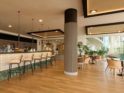 Área de bar moderna en hotel con mostrador, taburetes altos y asientos cómodos con luz natural.