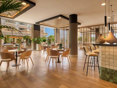 Área de salón moderna de hotel con suelo de madera, sillas claras, plantas y grandes ventanales.