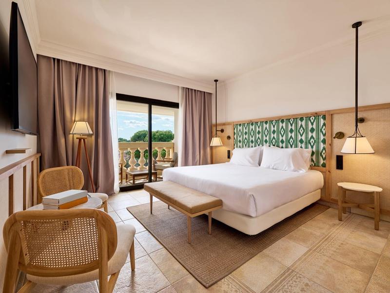 Jasny pokój hotelowy z łóżkiem podwójnym, częścią wypoczynkową i balkonem z widokiem na zielony krajobraz.