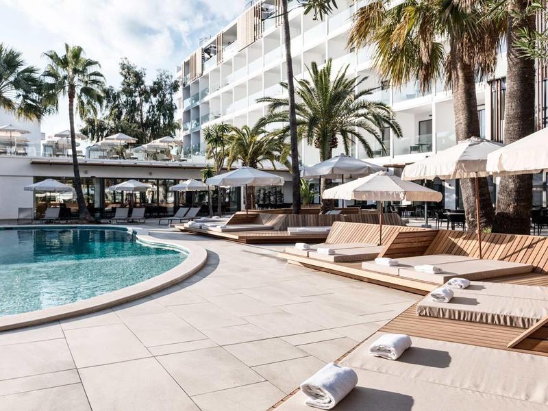 Area piscina di un hotel moderno con lettini, ombrelloni e palme in una giornata di sole.