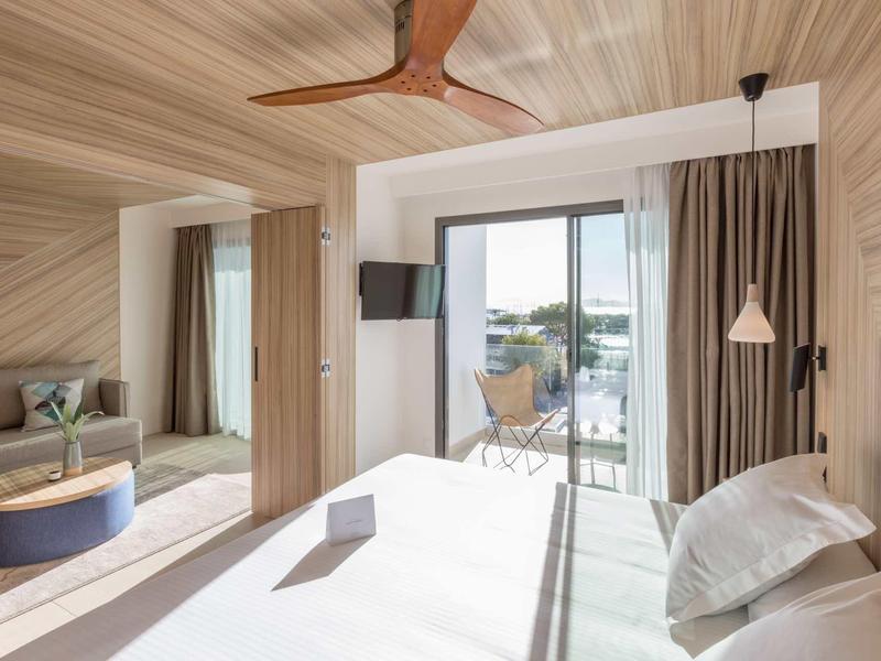 Camera d'hotel luminosa con letto grande, pareti in legno, ventilatore a soffitto e balcone con vista.