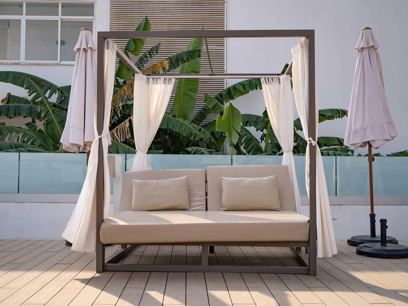 Divano doppio accogliente con baldacchino e tende bianche su una terrazza soleggiata accanto a ombrelloni chiusi.