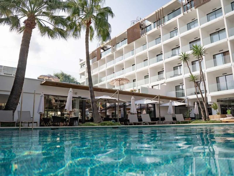 Edificio dell'hotel con piscina circondata da palme sotto un cielo limpido diurno.