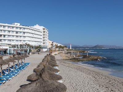 Strand mit Sonnenschirmen, Liegen und weißem Hotelgebäude am hellen Sand unter blauem Himmel.