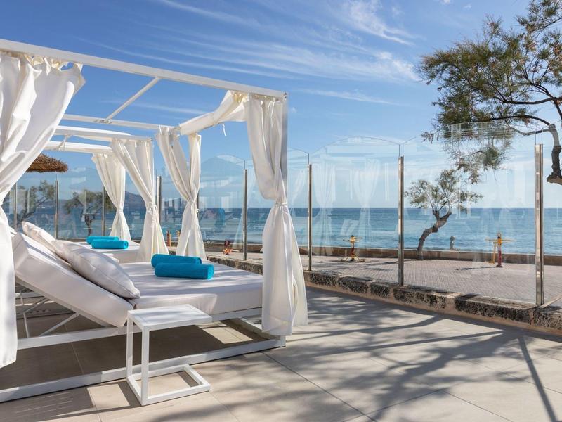 Weiße Himmelbett-Liege mit Vorhängen am sonnigen Meerufer unter blauem Himmel.