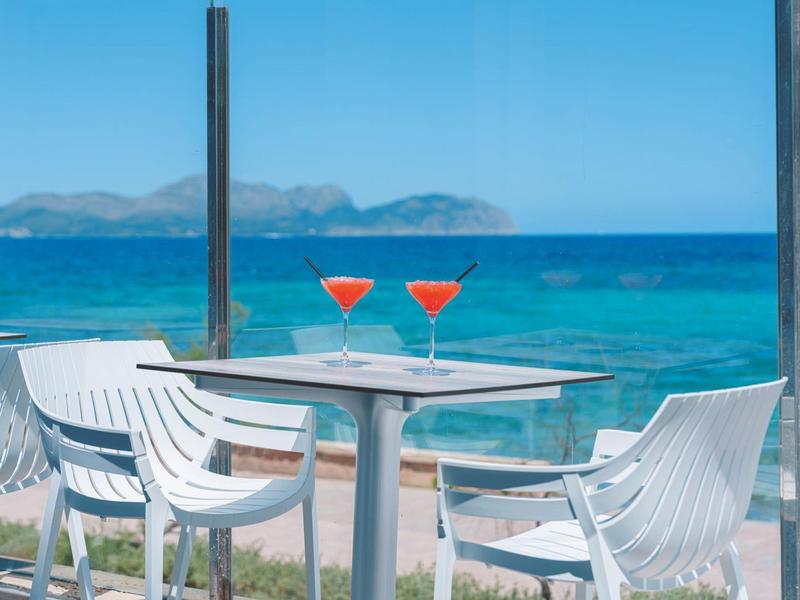 Terrasse mit weißen Stühlen und einem Tisch mit zwei roten Cocktails mit Blick auf das blaue Meer und entfernte Berge.