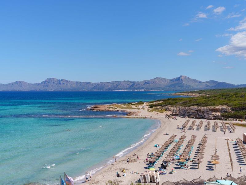 Strand mit klar blauem Wasser, Liegestühlen und Sonnenschirmen neben einer bewaldeten Küste unter blauem Himmel