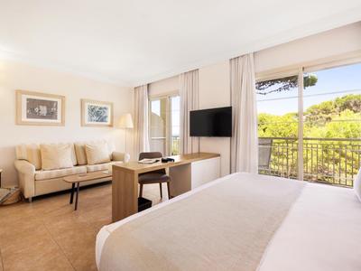 Chambre d'hôtel lumineuse avec lit, canapé, bureau et balcon avec vue sur des arbres verts.