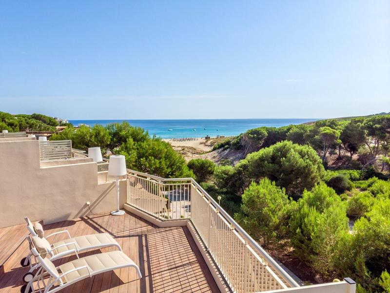 Balcon avec terrasse en bois et chaises avec vue sur la mer et les dunes vertes sous un ciel bleu.