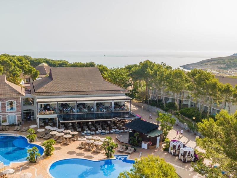 Hôtel avec grandes piscines, terrasse ensoleillée et vue sur la mer derrière des arbres.