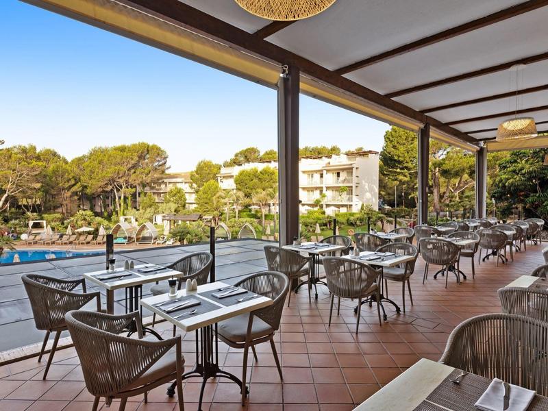 Terrasse extérieure couverte avec tables surplombant la piscine et l'hôtel sous un ciel clair.