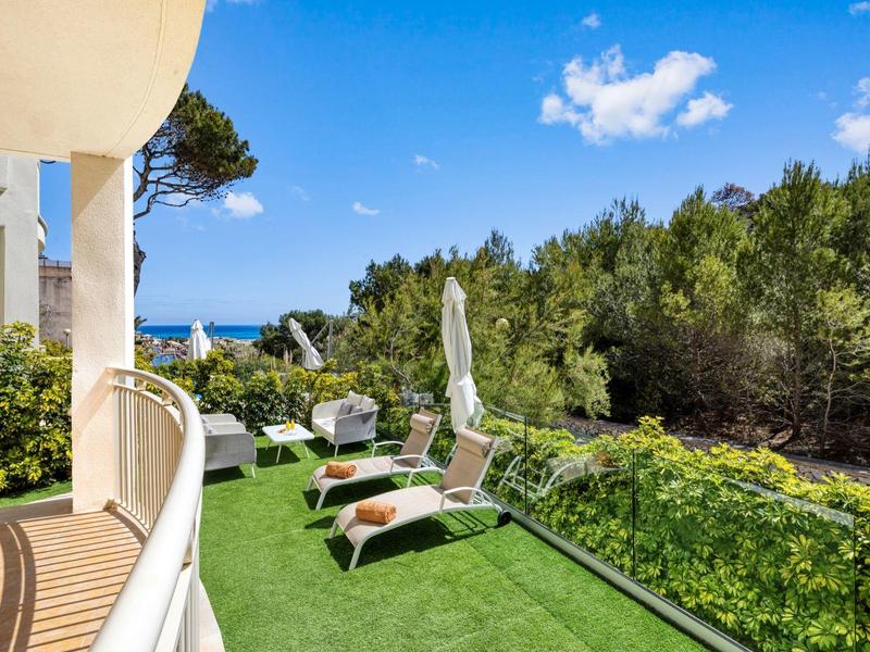 Balcon avec chaises longues et parasol, vue sur la forêt et la mer sous un ciel bleu.