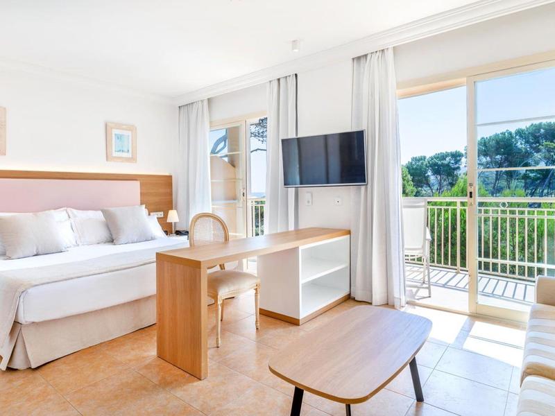 Chambre d'hôtel lumineuse avec lit double, canapé et balcon donnant sur la verdure.