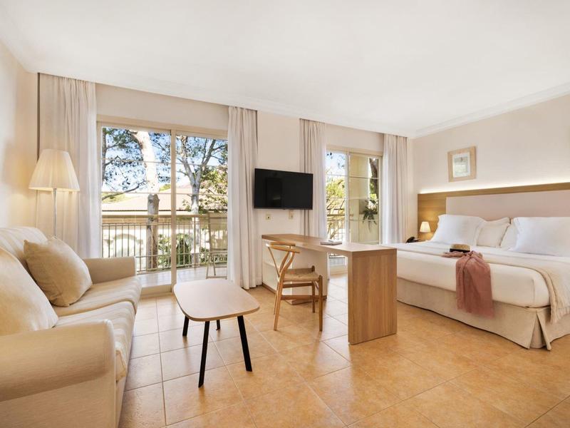 Chambre d'hôtel lumineuse avec lit double, canapé, TV et vue sur balcon avec arbres.