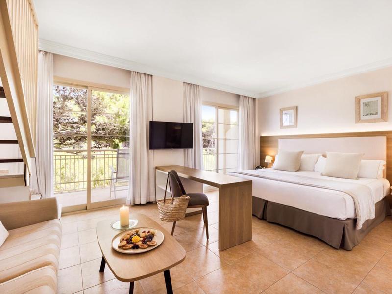 Chambre d'hôtel lumineuse avec lit double, canapé, table et vue sur jardin par grandes fenêtres.