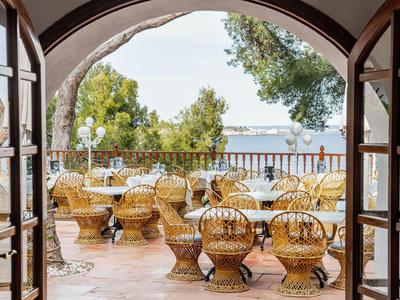 View through an archway to a terrace with round chairs and tables overlooking the sea.