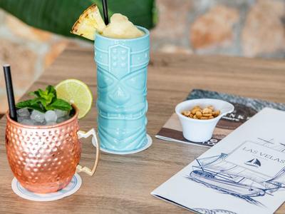 Two colorful cocktails on a wooden table with a sailboat menu and small snacks.