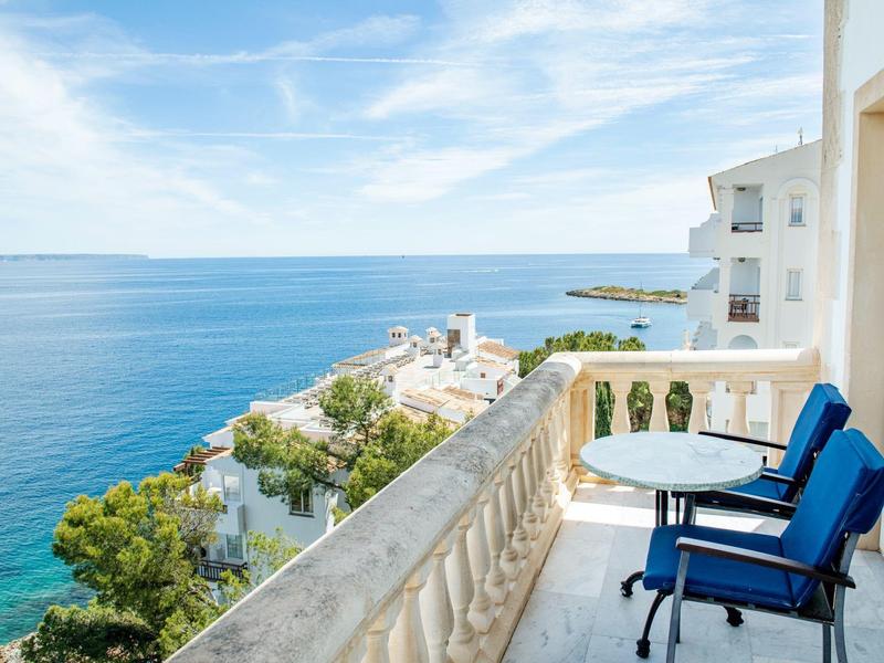 Balcony with blue cushioned chairs and table overlooking the sea under clear sky.