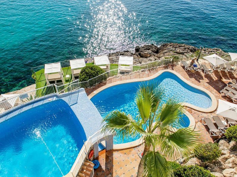 Two pools with sun loungers on a terrace by the sea and a palm tree in the foreground.