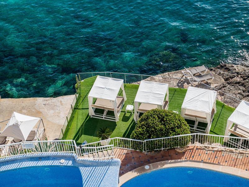 View of a pool area by the sea with white cabanas and clear blue water.
