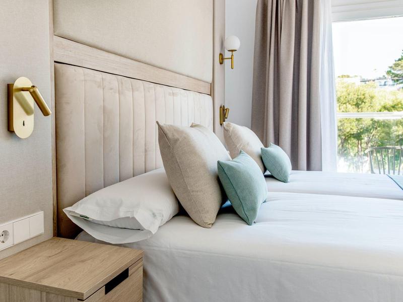 Modern hotel room with two beds, beige headboards, multiple pillows, and window view outside.