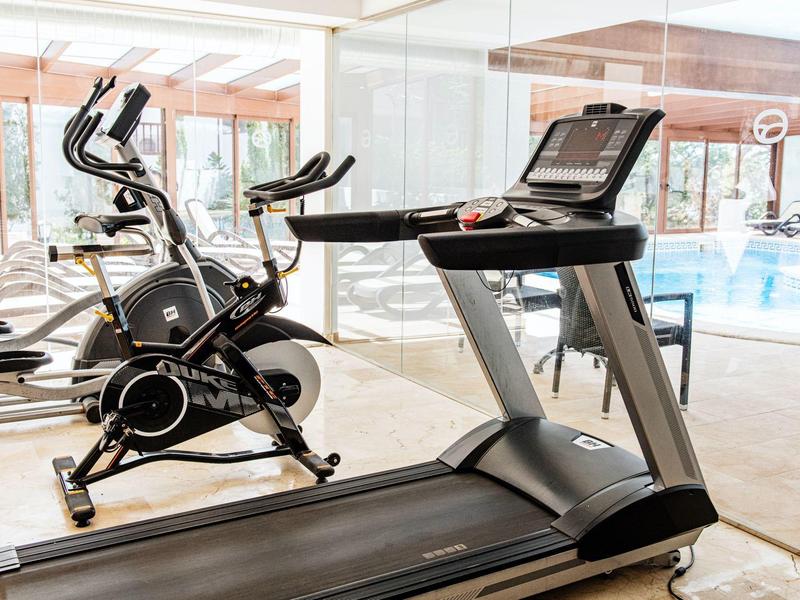 Modern fitness equipment like treadmill and exercise bike in a bright gym with pool view.