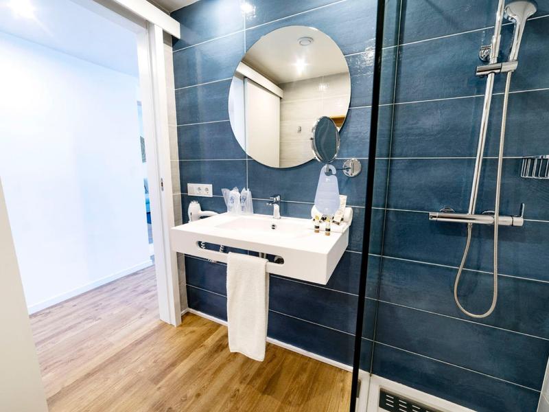 Helles Badezimmer mit runder Spiegel, weißem Waschbecken, dunkler Duschwand und Holzboden.