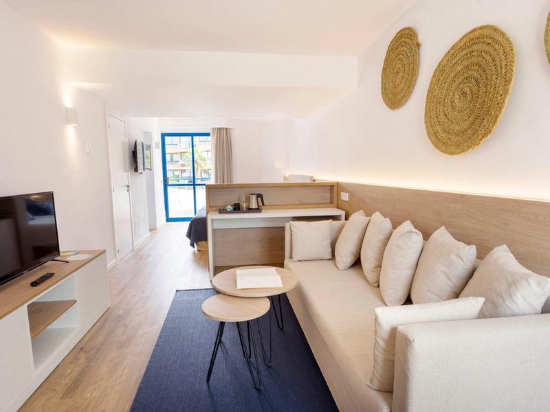 Helles Wohnzimmer mit beiger Couch, Holzboden, blauem Teppich und TV auf weißem Regal.