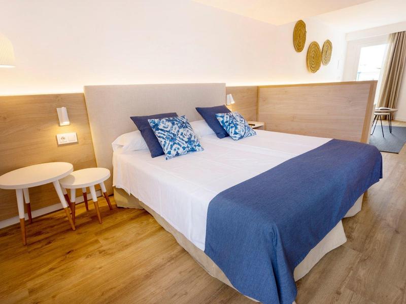 Helles Schlafzimmer mit Doppelbett, blauen Kissen und Decke, Holzfußboden und weißem Beistelltisch.