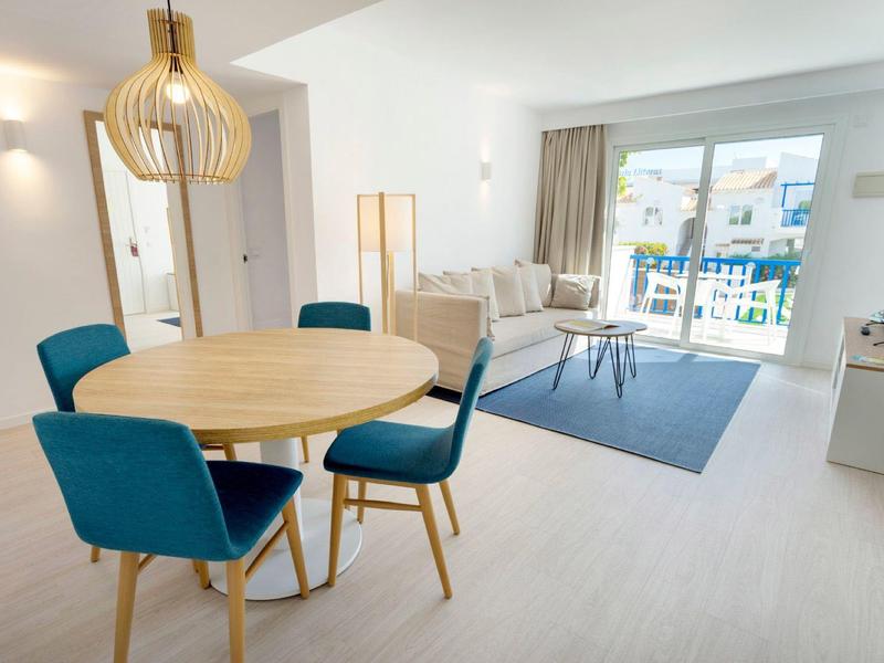 Helles Wohnzimmer mit runder Holztisch, blauen Stühlen, Sofa, großen Fenstern und modernem Lampenschirm.